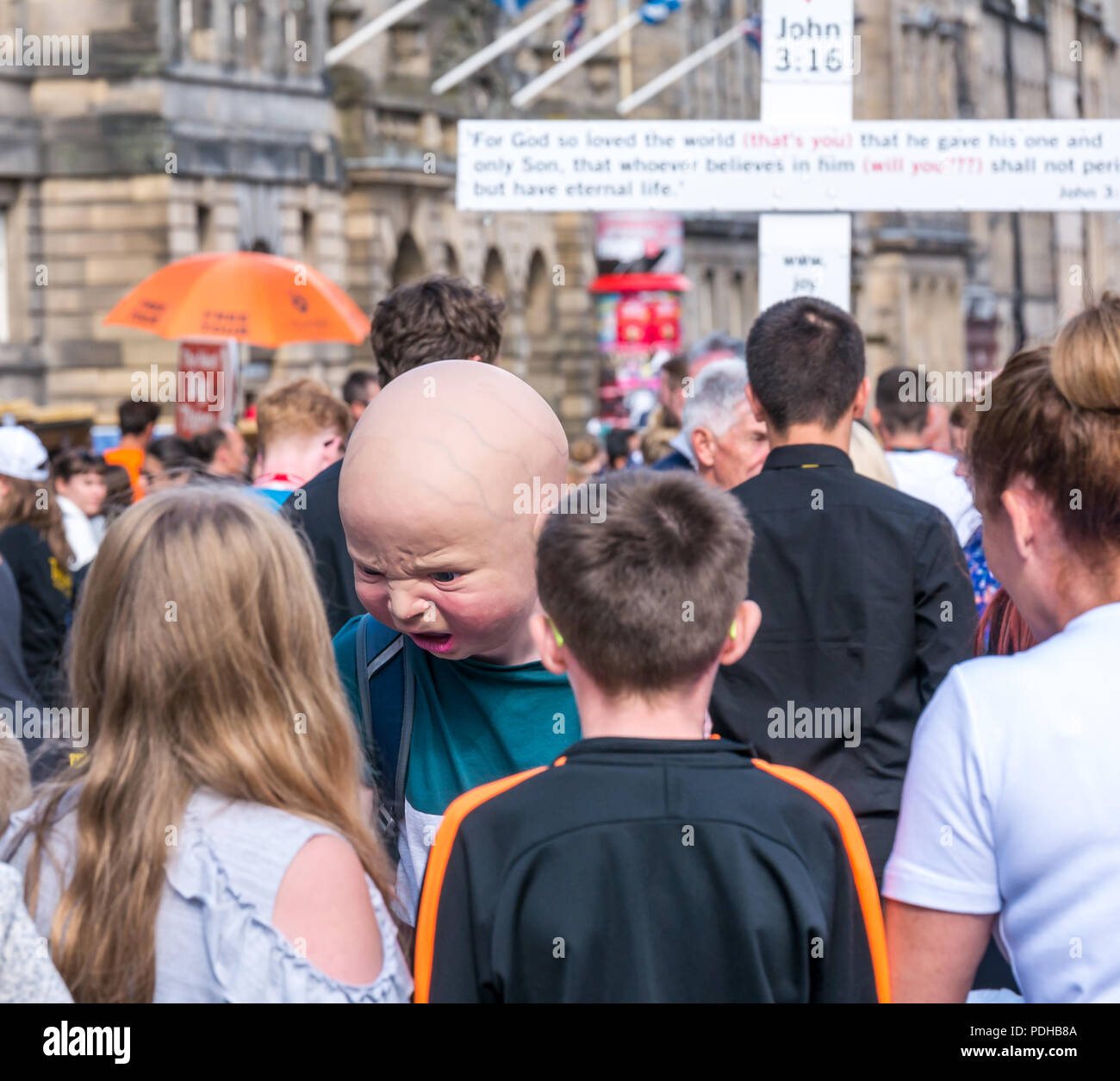 Edinburgh, Scotland, UK. 9th August 2018. Edinburgh Fringe Festival, Royal Mile, Edinburgh, Scotland, United Kingdom. On a sunny festival day the Virgin Money sponsored street festival is packed with people and fringe performers. A Fringe performer wearing a gruesome baby head interacts with pedestrians handing out flyers Stock Photo