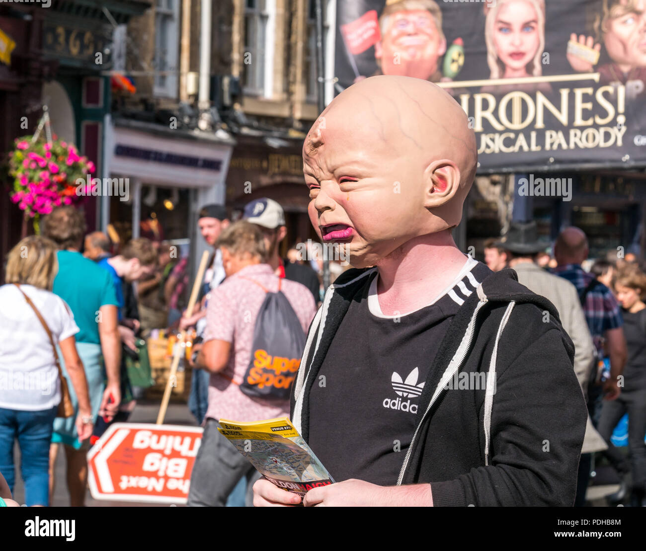 Edinburgh, Scotland, UK. 9th August 2018. Edinburgh Fringe Festival, Royal Mile, Edinburgh, Scotland, United Kingdom. On a sunny festival day the Virgin Money sponsored street festival is packed with people and fringe performers. A Fringe performer wearing a gruesome baby head interacts with pedestrians handing out flyers Stock Photo