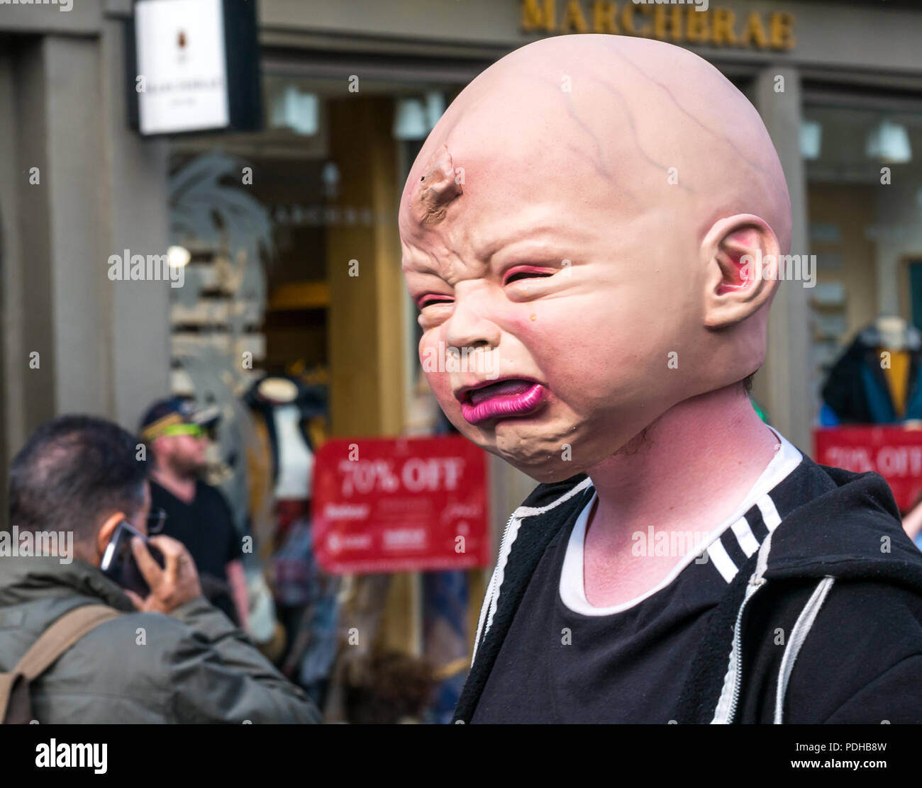 Edinburgh, Scotland, UK. 9th August 2018. Edinburgh Fringe Festival, Royal Mile, Edinburgh, Scotland, United Kingdom. On a sunny festival day the Virgin Money sponsored street festival is packed with people and fringe performers. A Fringe performer wearing a gruesome baby head interacts with pedestrians handing out flyers Stock Photo