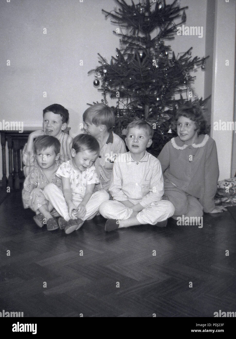 1960s, excited young chiildren sit on a wooden floor of a room at Christmas in their pyjamas and slippers infront of a decorated Xmas tree, awaiting their presents, England, UK. Stock Photo