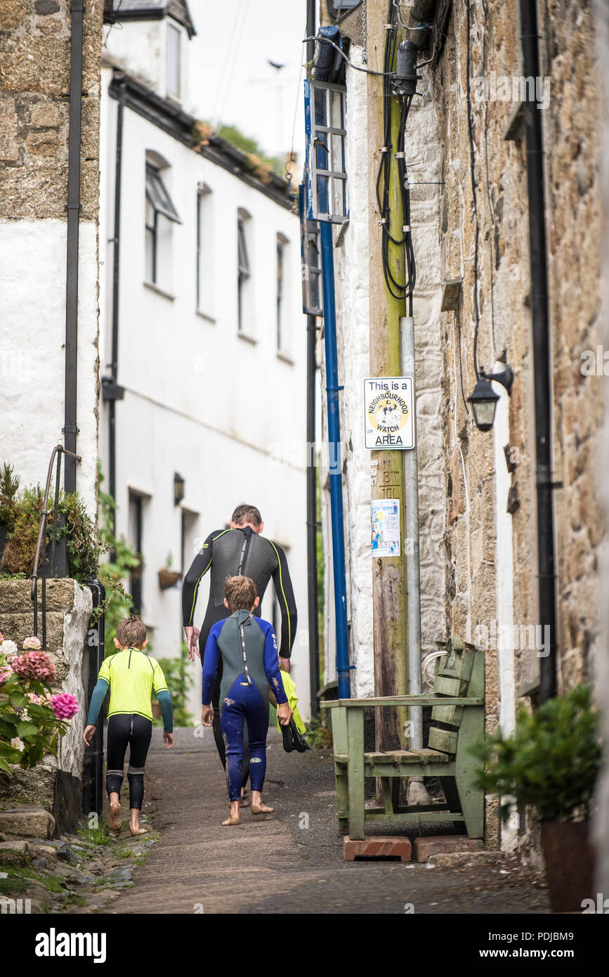 A man and two childrn wearing wetsuits walking up Duck street in Mousehole in Cornwall. Stock Photo