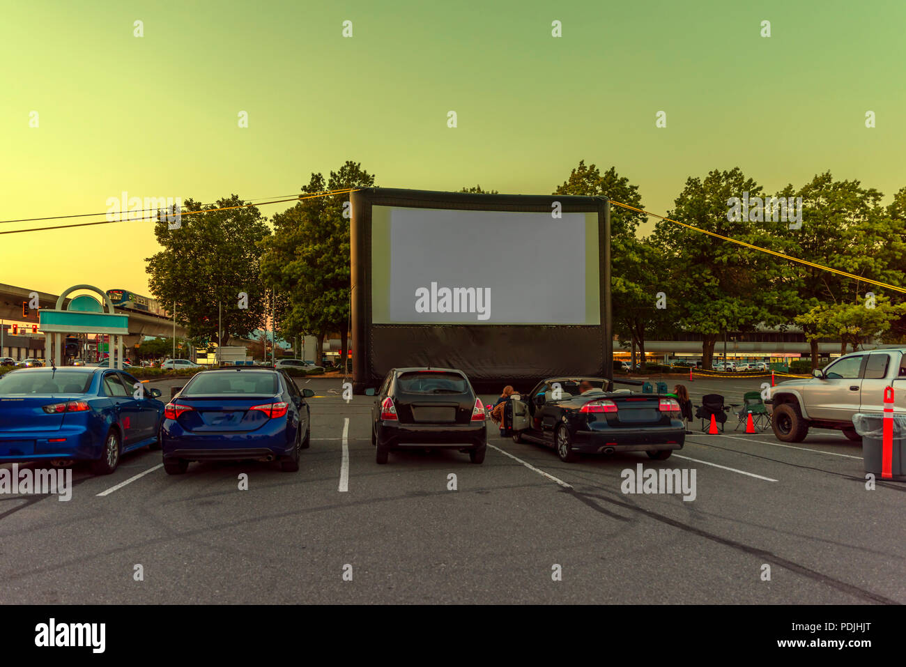Spectators at a car parking lot with cars, an inflatable screen of the summer cinema, waiting for a movie. A city train passes through a viaduct. Stock Photo