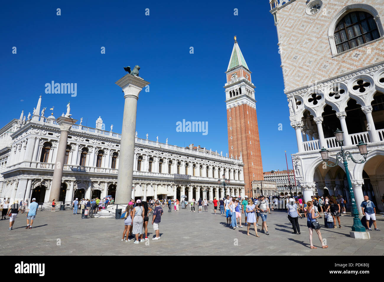 San Marco bell tower, National Marciana library facade, lion statue and people in the square, clear blue sky in Venice Stock Photo