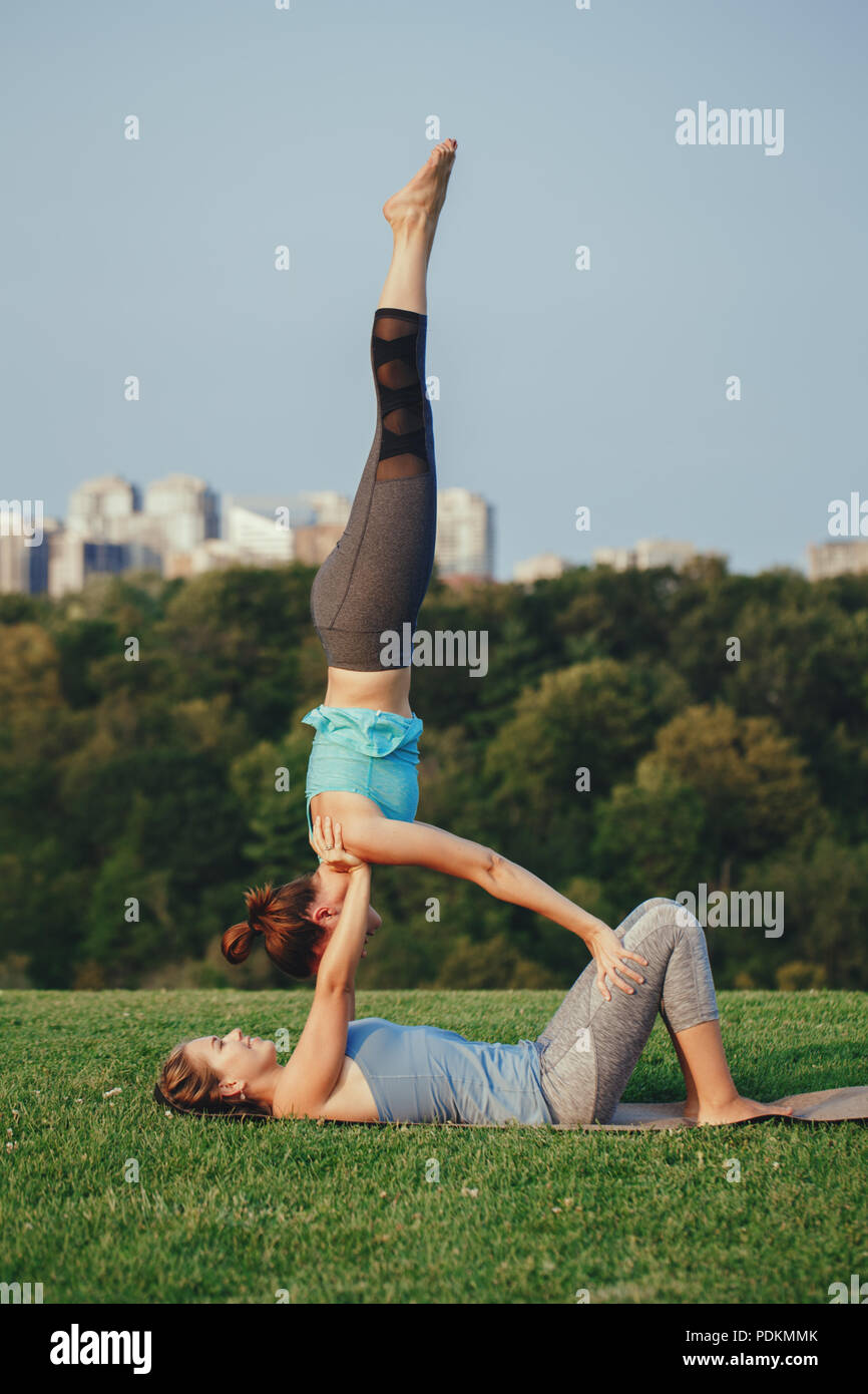 Two Caucasian women yogi doing shoulder stand candlestick acro yoga pose. Women doing stretching workout in park outdoors at sunset. Healthy lifestyle Stock Photo