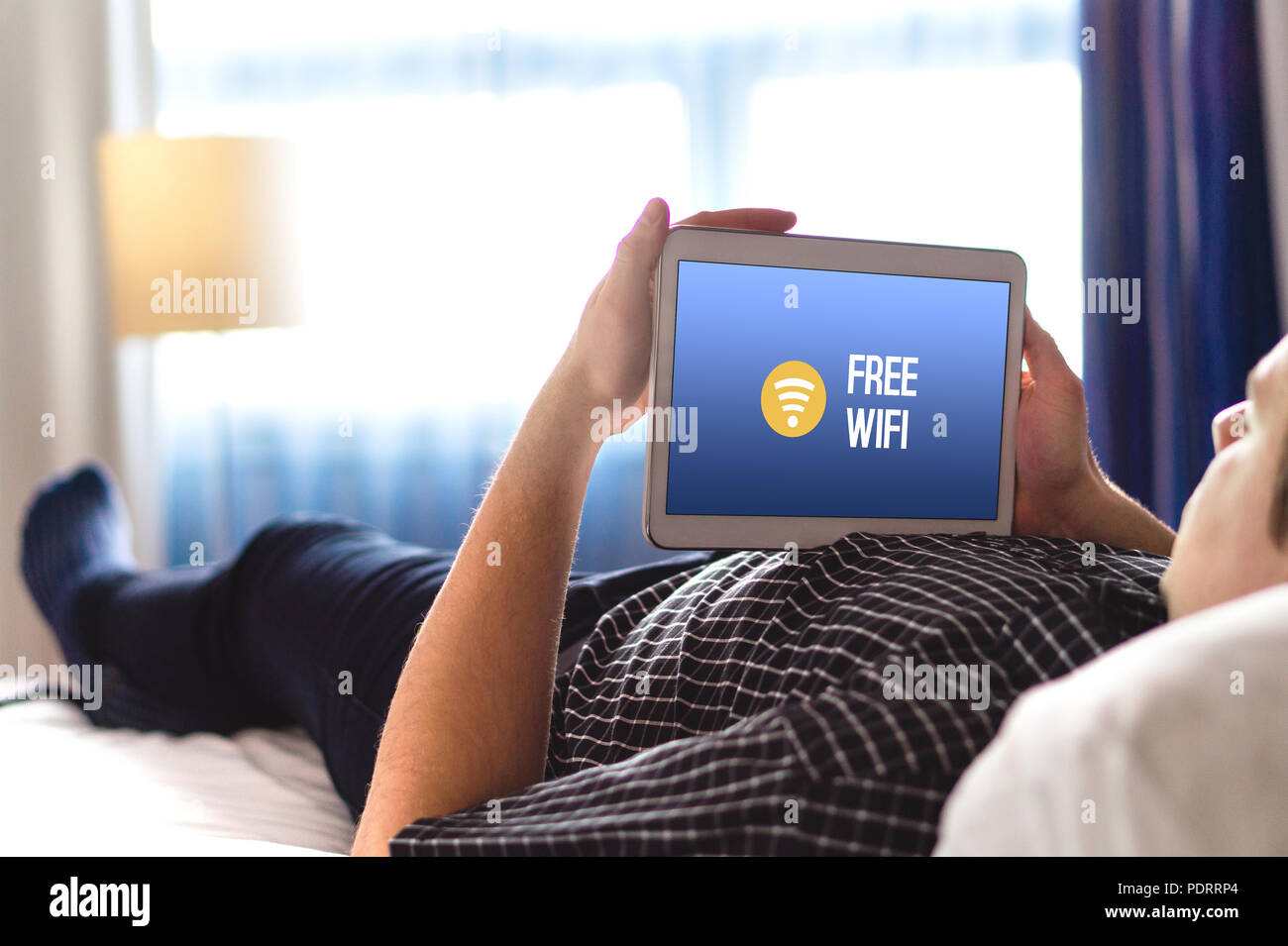 Man using free hotel wifi with tablet. Lying in hotel room bed and browsing internet. Public access online and connection available for customers. Stock Photo