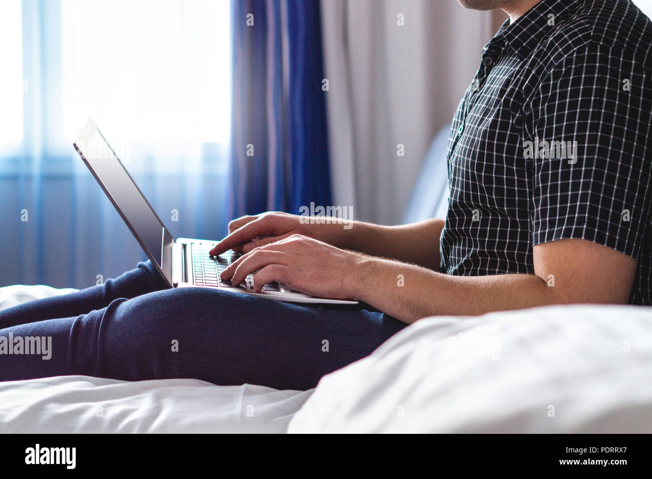 Man using laptop in hotel room or home bedroom. Guy lying on bed and typing with notebook computer. Person in the morning with modern device. Stock Photo