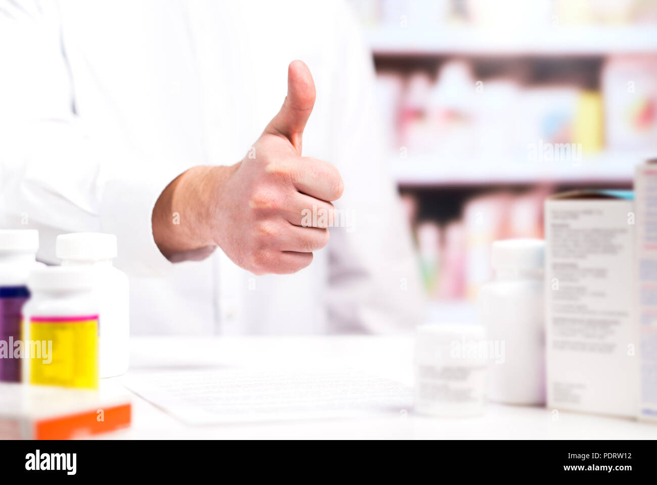 Happy pharmacist showing thumbs up at pharmacy counter full of medicine. Drugstore shelves in the background. Positive and cheerful druggist. Stock Photo