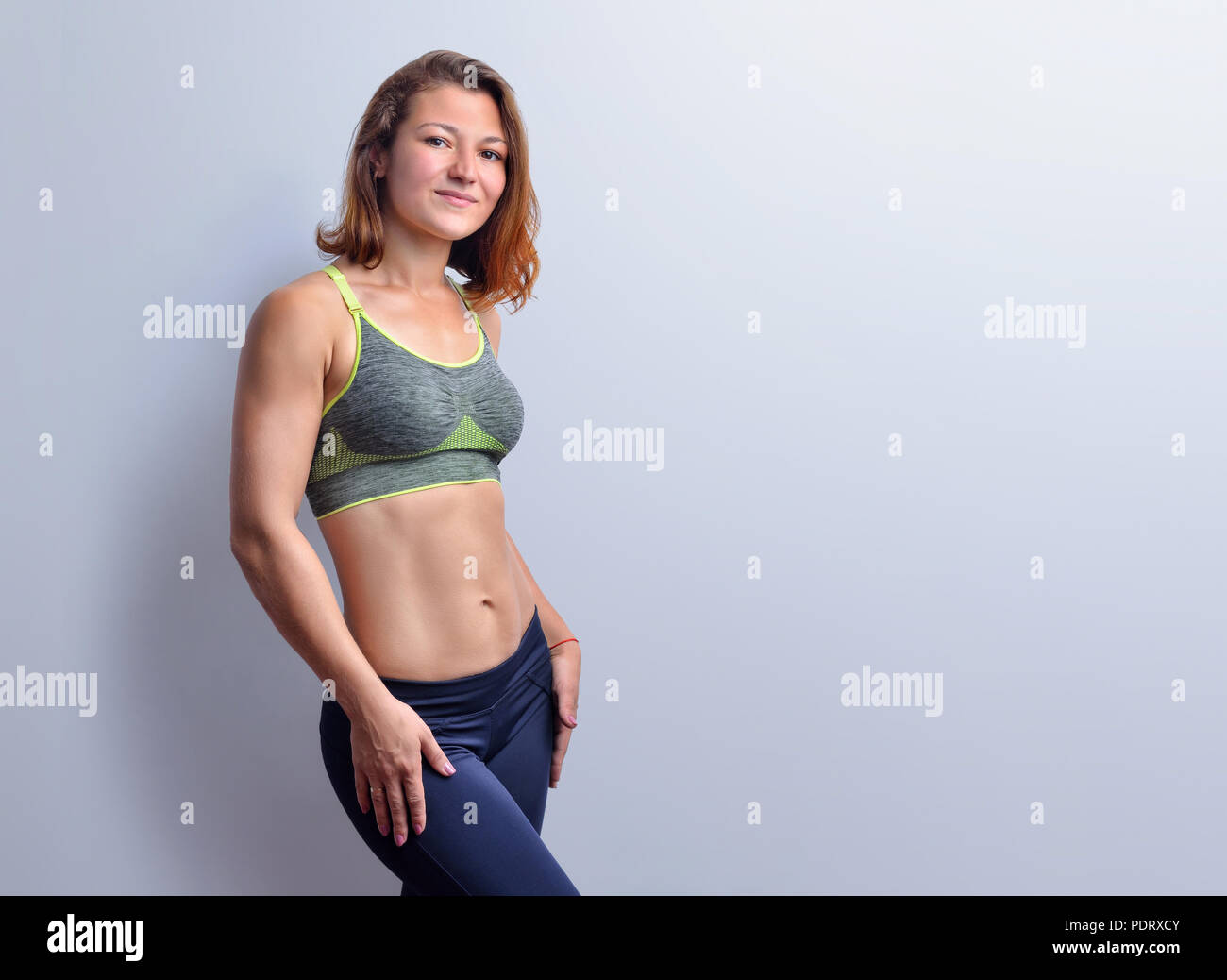 Smiling female fitness coach in grey top and black leggings with slim abdominal muscles  is posing on the white background, front view. Stock Photo