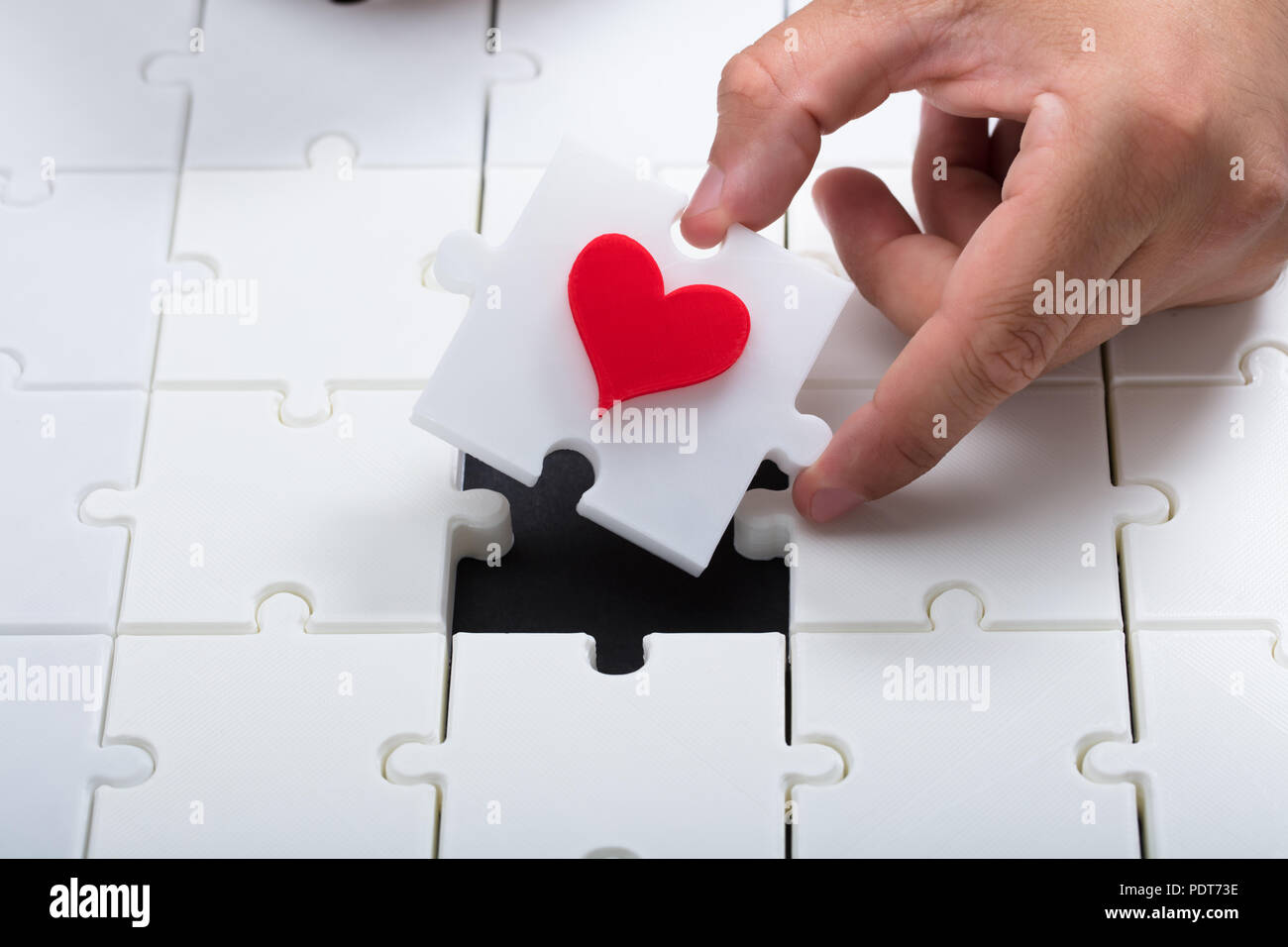 Person's hand placing last piece with red heart into white jigsaw puzzle Stock Photo
