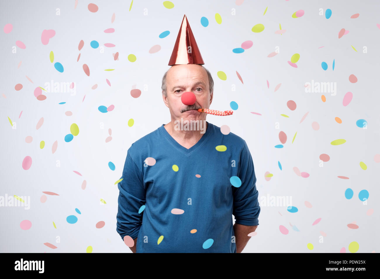 Unhappy birthday senior man is sad and disappointed because nobody came to celebrate his anniversary. Blowing party horn, funny cap and red clown nose Stock Photo