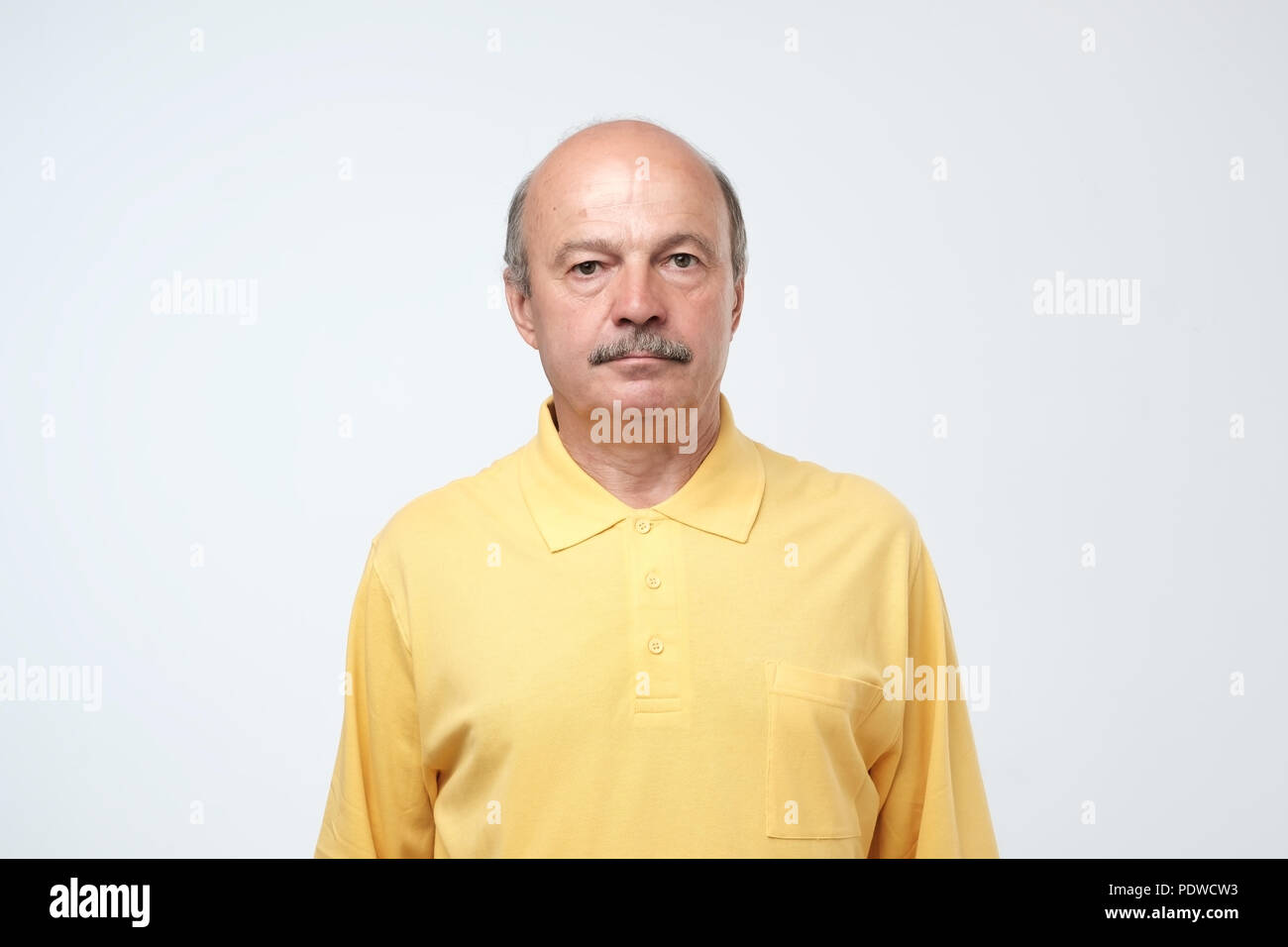Elderly man emotions, portrait of serious senior caucasian man in yellow t-shirt looking at camera against gray wall Stock Photo