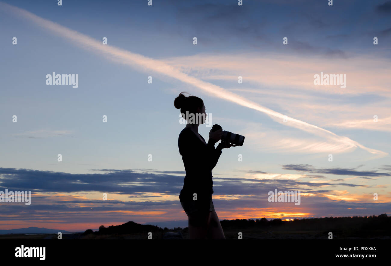 A girl silhouetted against a sunset sky, taking a photo with a DSLR. Stock Photo