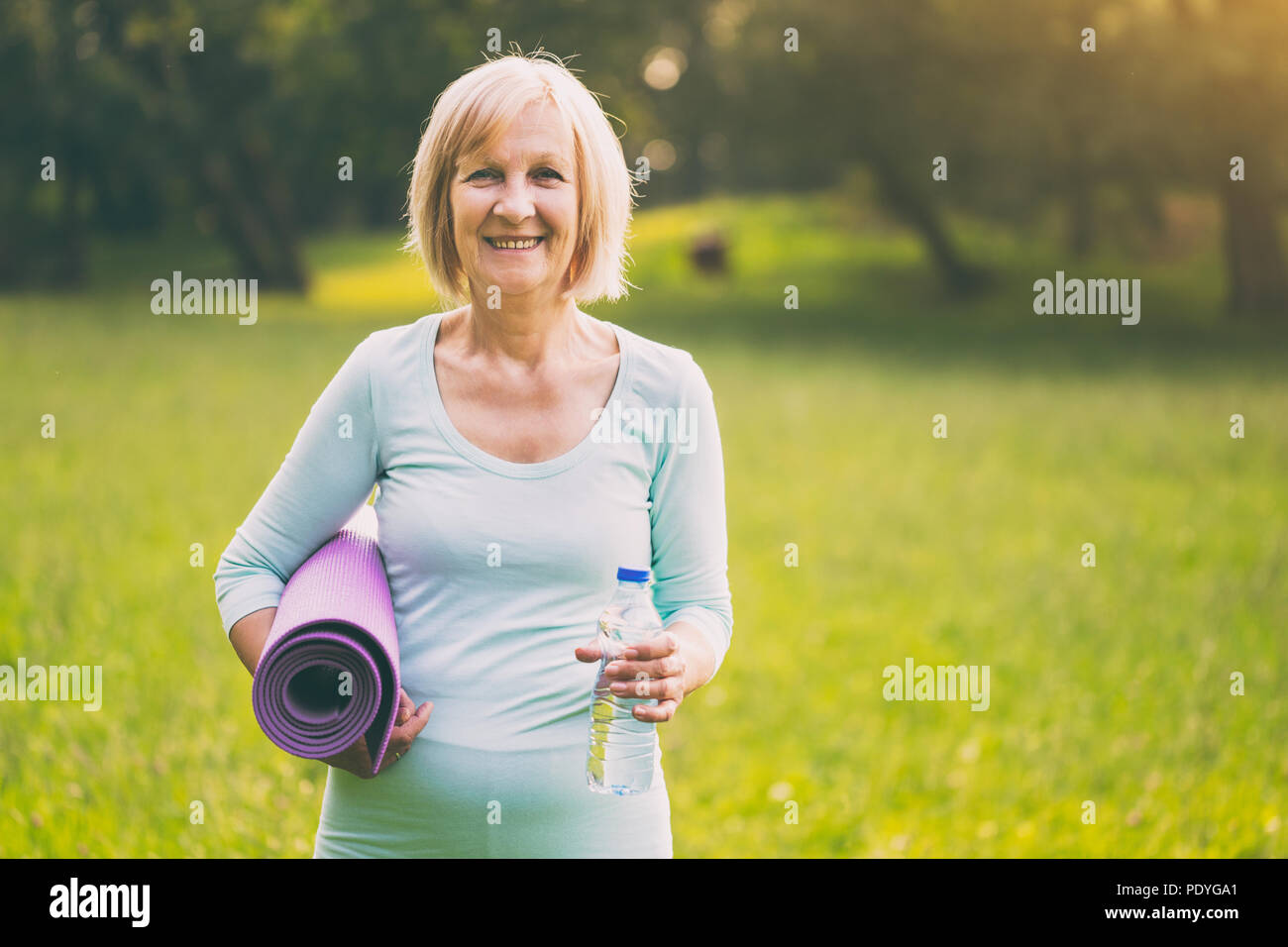 Portrait of sporty senior woman holding bottle of water and mat in the nature.Image is intentionally toned. Stock Photo