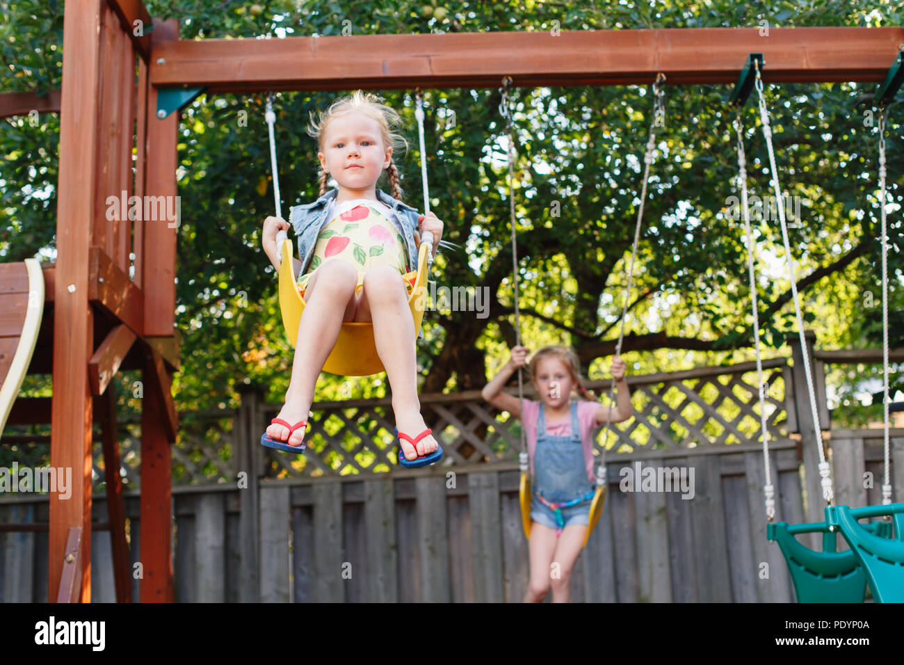 Portrait of two happy smiling little girls on swing on backyard playground outside on summer day. Sisters having fun together, leisure time. Happy chi Stock Photo