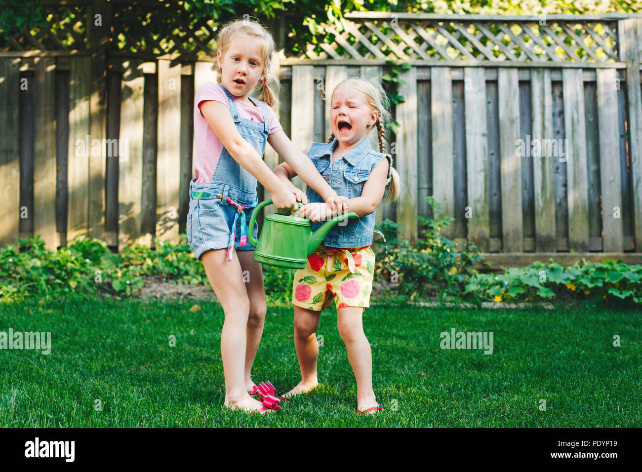 Portrait of  two little girls  sisters having fight on home backyard. Friends girls share watering pot can. Lifestyle family moment of siblings quarre Stock Photo