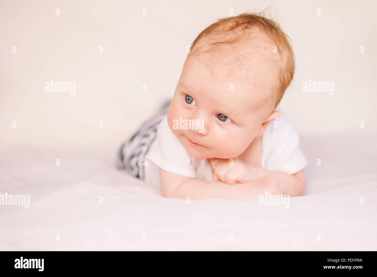 Closeup portrait of adorable funny white Caucasian baby with blue grey eyes lying on tummy belly on bed. Aware cute newborn on white light background  Stock Photo