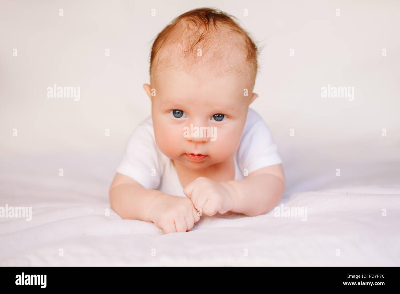 Closeup portrait of adorable funny white Caucasian baby with blue grey eyes lying on tummy belly on bed. Aware cute newborn on white light background  Stock Photo