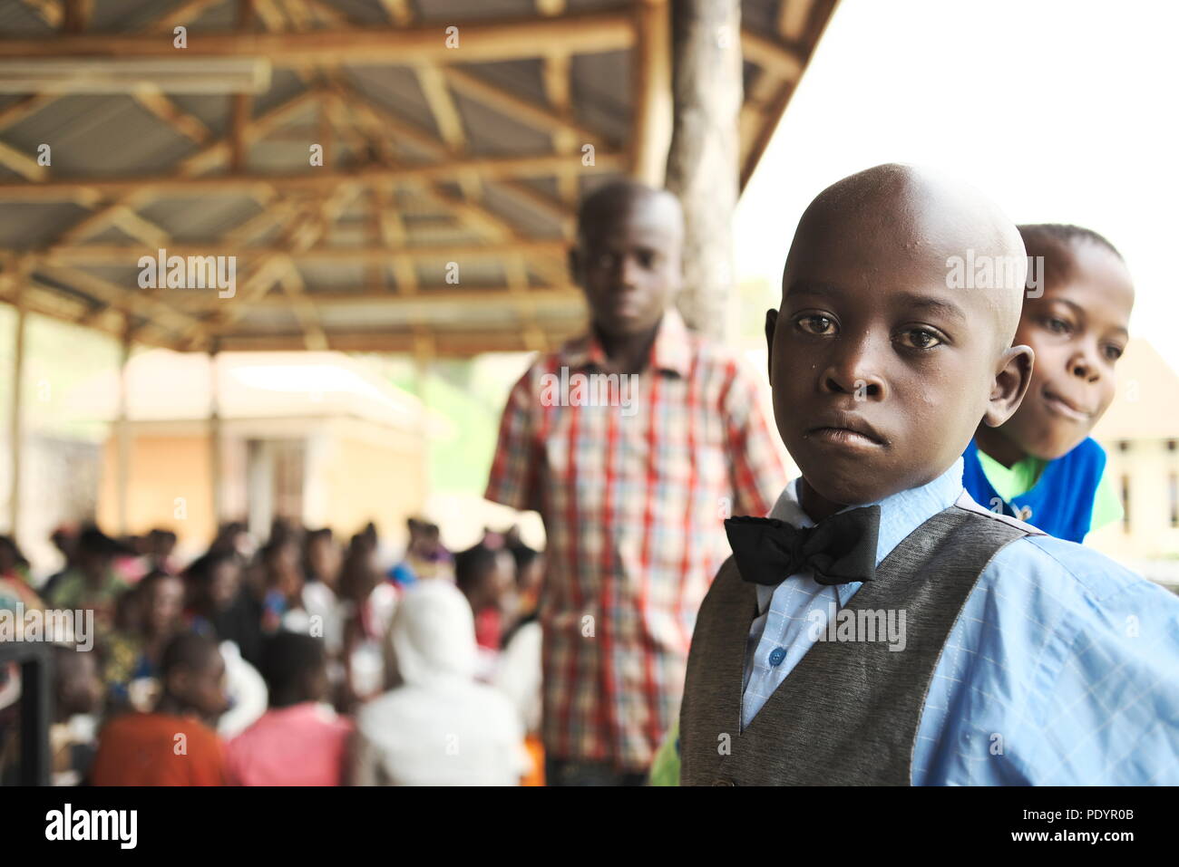 Young Ugandan boy stands in his Sunday best, staring at the camera with a group of other young African children waiting for hospital attention Stock Photo