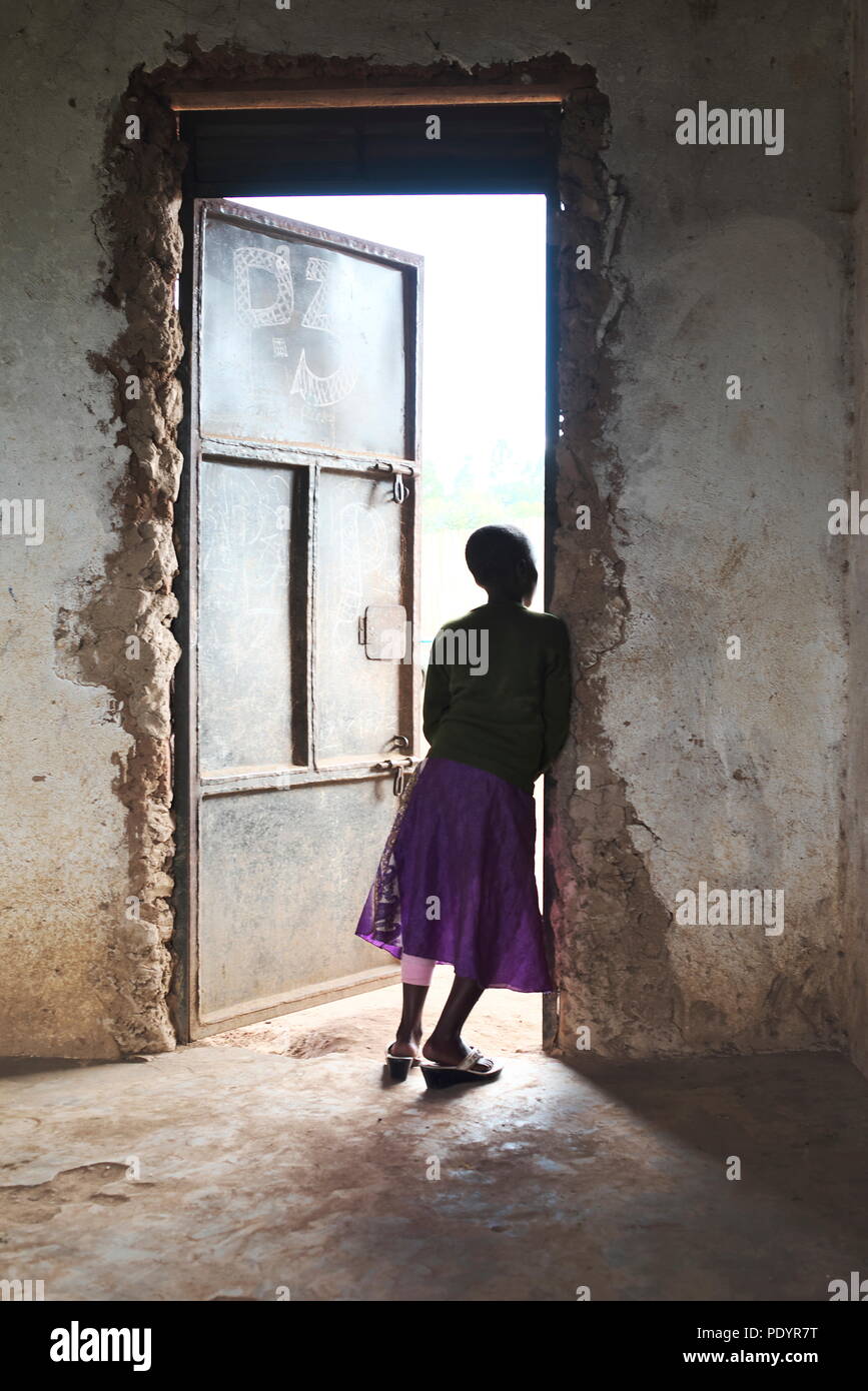 Young Ugandan girl stands in the doorway of her school classrom looking out at other pupils, wearing a bright coloured skirt Stock Photo