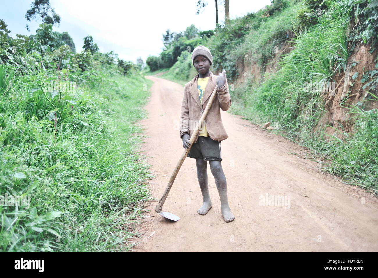 Young Ugandan boy stands in the middle of a dirt road holding farm tools, barefoot, looking at the camera Stock Photo