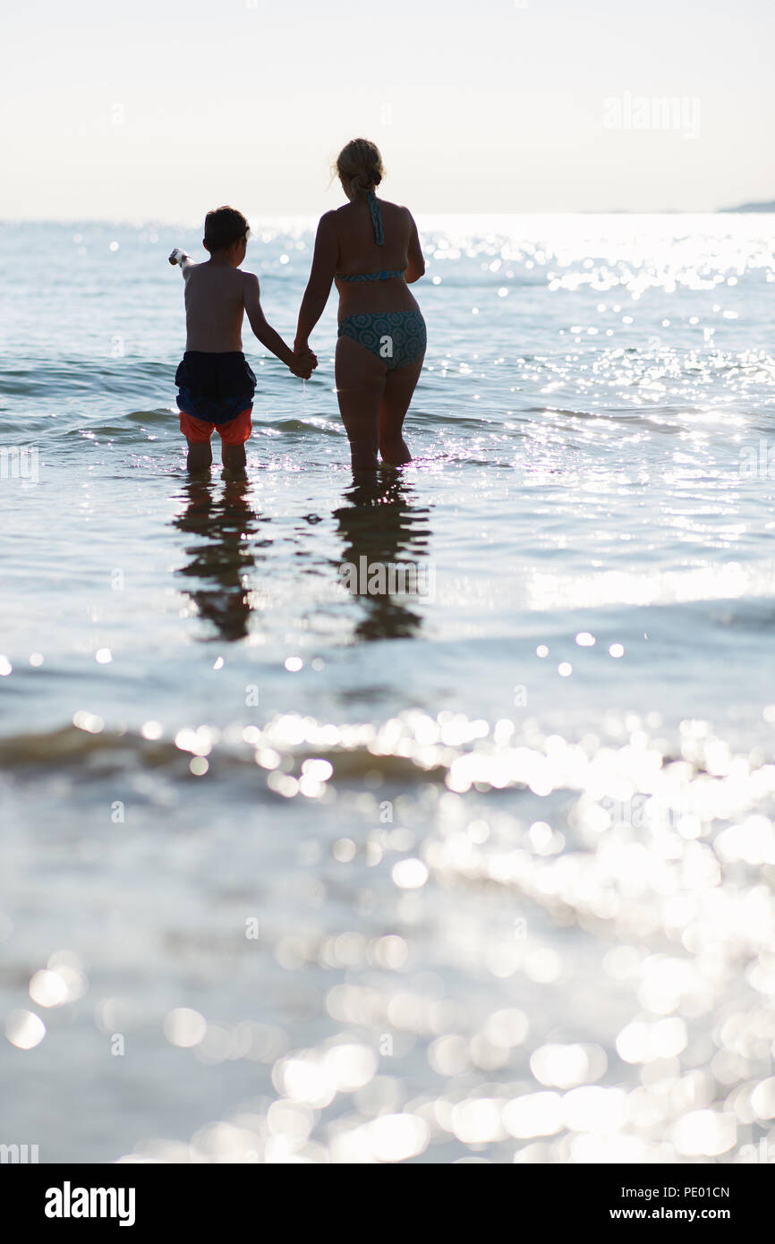 Mother and son having fun jumping the waves of the sea in the early evening whilst the sun is setting and is glistening on the pure blue water Stock Photo