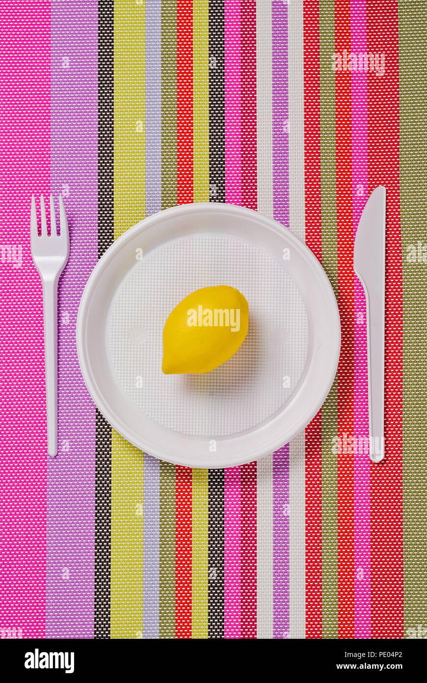 White plastic utensils, fork, plate and knife on a colored striped napkin. And yellow lemon in a plate. Stock Photo