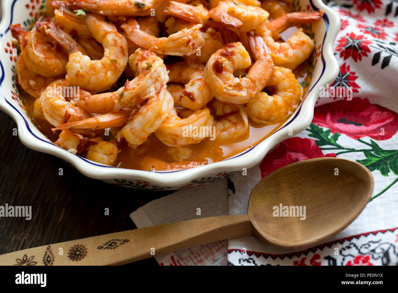 Juicy appetizing Shrimps cooked and baked in sauce are displayed on a wooden table in a dish with ornament located next to a towel with colorful ornam Stock Photo