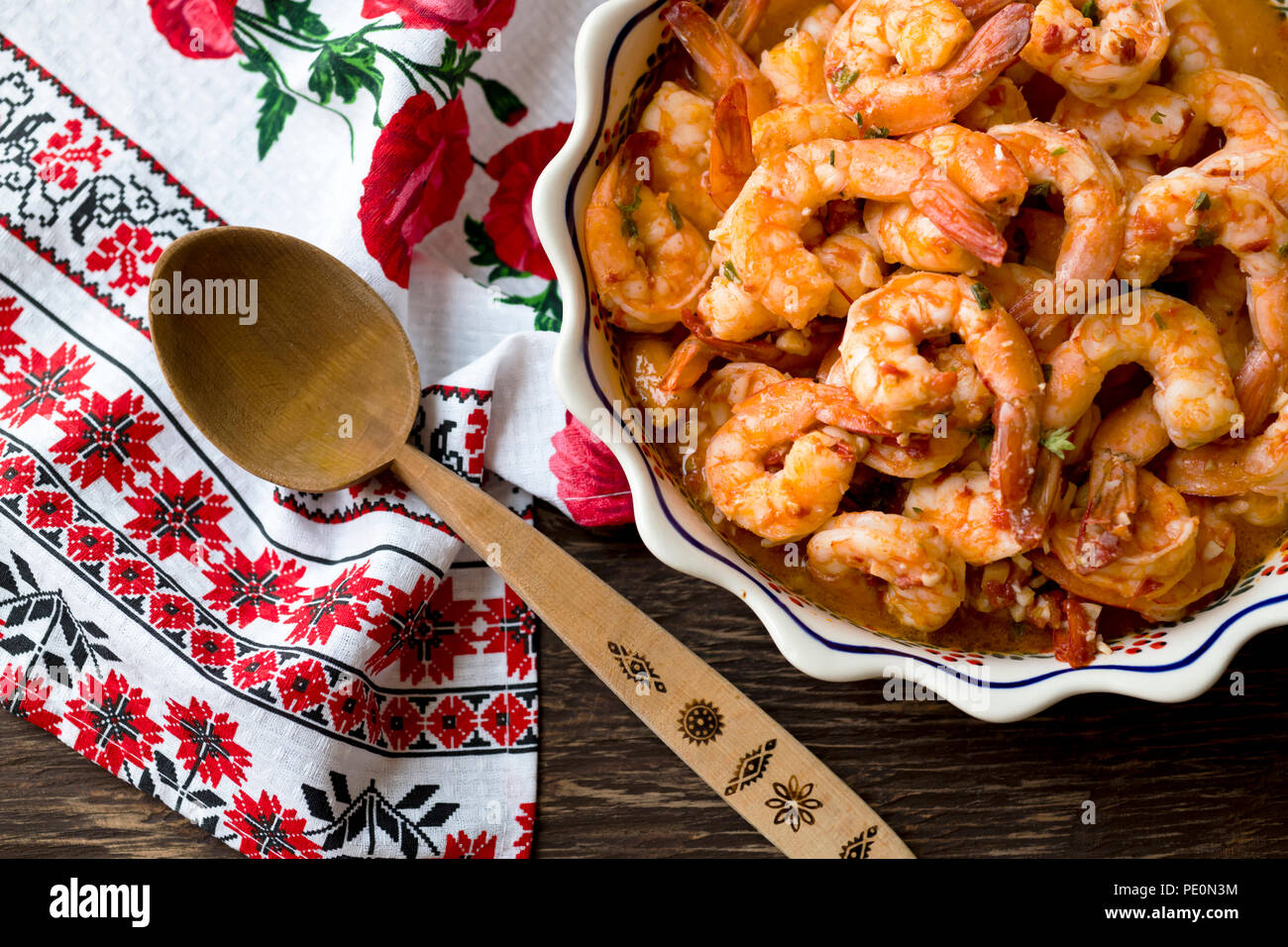 Juicy appetizing Shrimps cooked and baked in sauce are displayed on a wooden table in a dish with ornament located next to a towel with colorful ornam Stock Photo