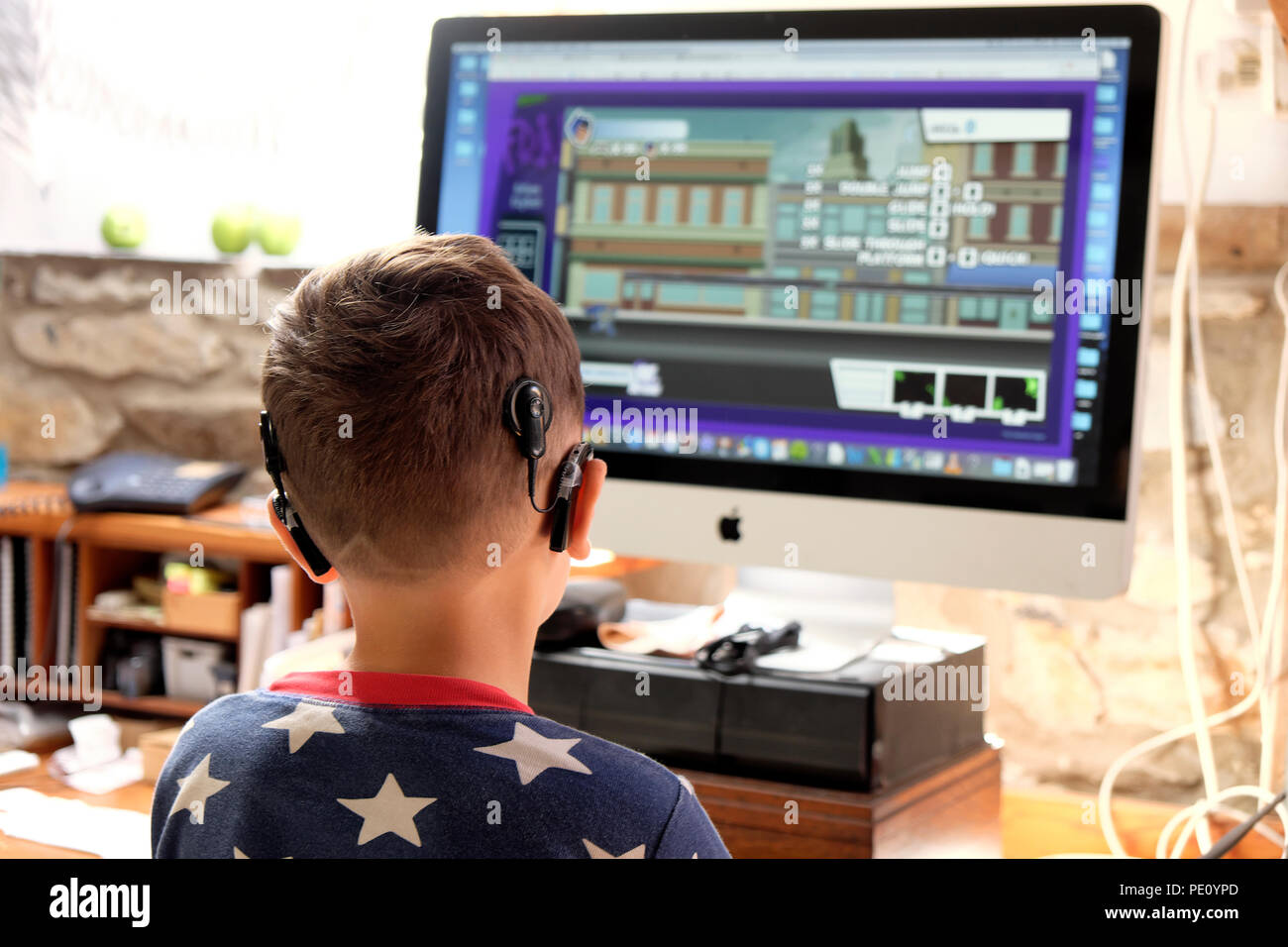 Rear back view of boy child wearing cochlear implant digital technology  looking at screen playing a computer game at home in the UK    KATHY DEWITT Stock Photo