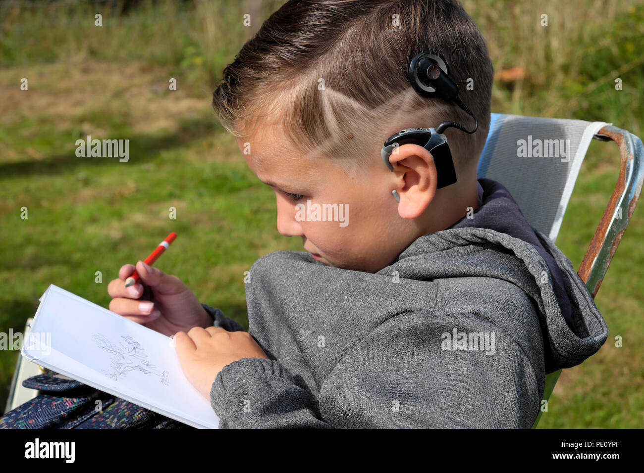 Side profile view of boy child 8 with cool star haircut sitting in garden drawing in a sketch book wearing cochlear implants device UK  KATHY DEWITT Stock Photo