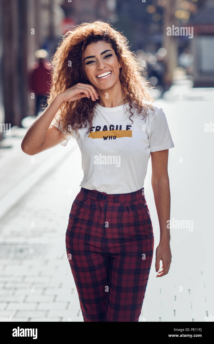 Smiling young arabic woman with black curly hairstyle. Arab girl in casual  clothes in the street. Happy female wearing white t-shirt and checked pants  Stock Photo - Alamy