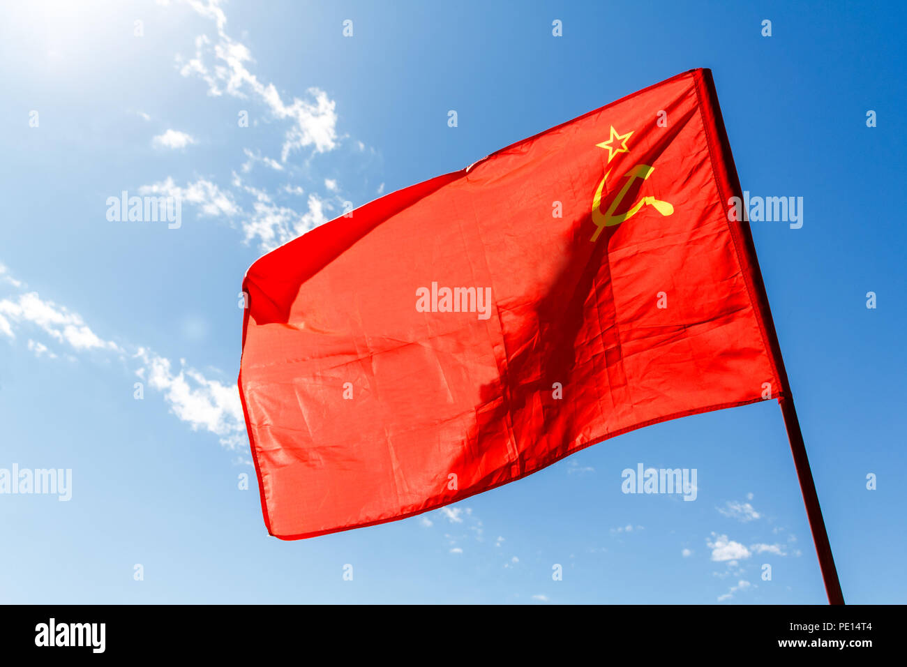 Red communist flag of the USSR against the blue sky with white clouds and sun. Stock Photo