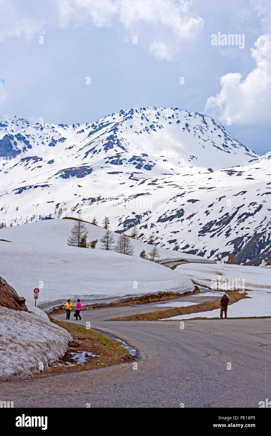 Pedestrians on road from summit of Simplon Pass to Ferienlager Kulm hotel from Hotel Simplon-Blick in early May.  Snow clad mountain in background Stock Photo