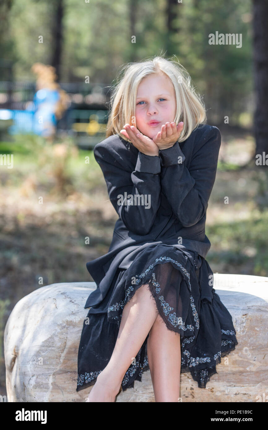 Attractive young girl, blonde, fashionably dressed in black outfit,, sitting outdoors on large rock, blowing kiss at camera. Stock Photo