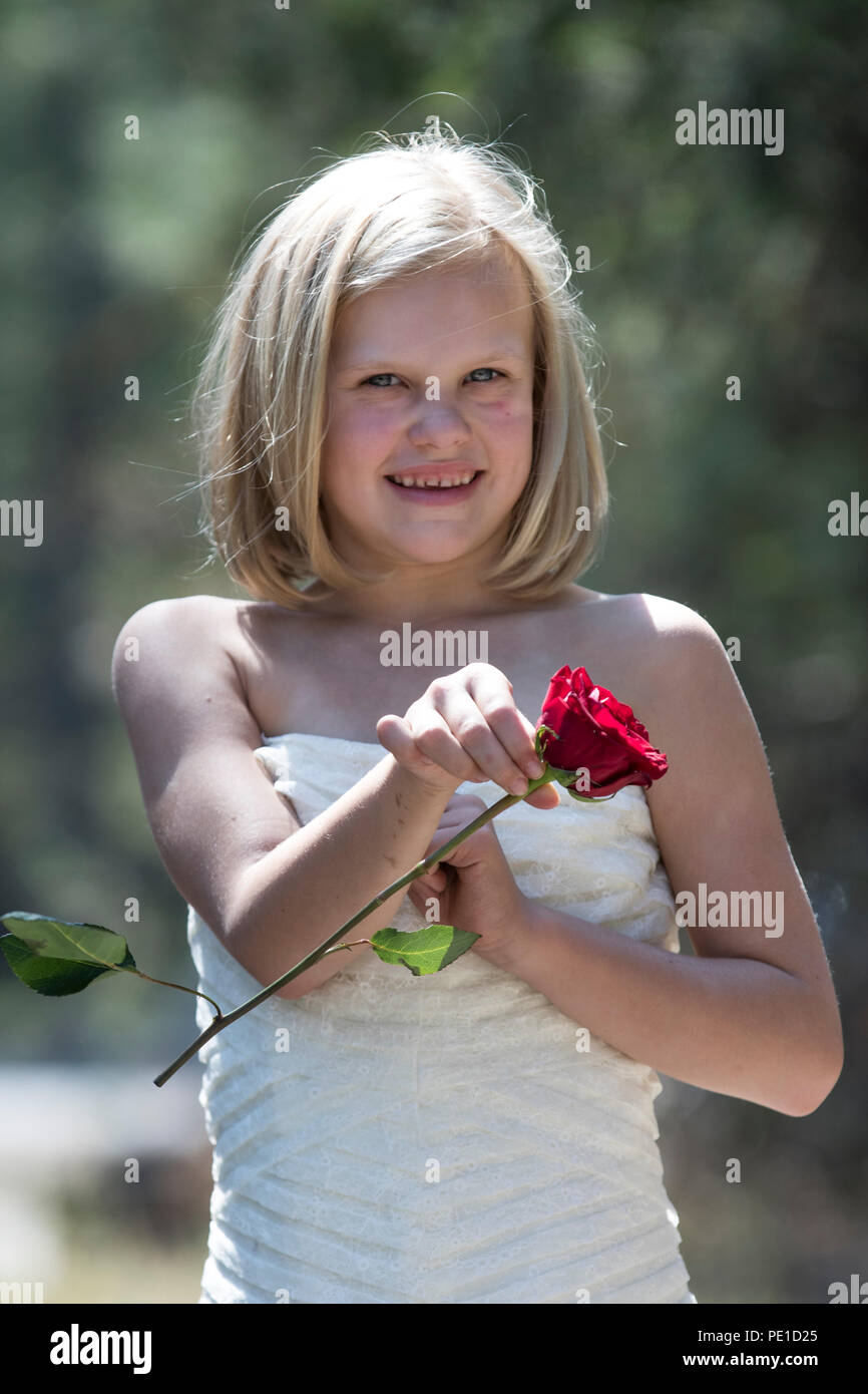 Fantasy, 8-9 year attractie blonde, wearing aunt's wedding dress . Standing outdoors, holding a red rose,  Smiling at camera. Stock Photo