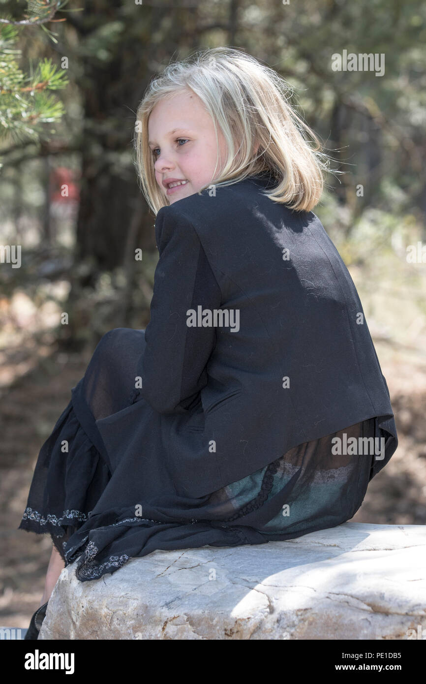 Attractive young girl, blonde, fashionably dressed in black outfit,, sitting outdoors on large rock, looking back at camera. Stock Photo