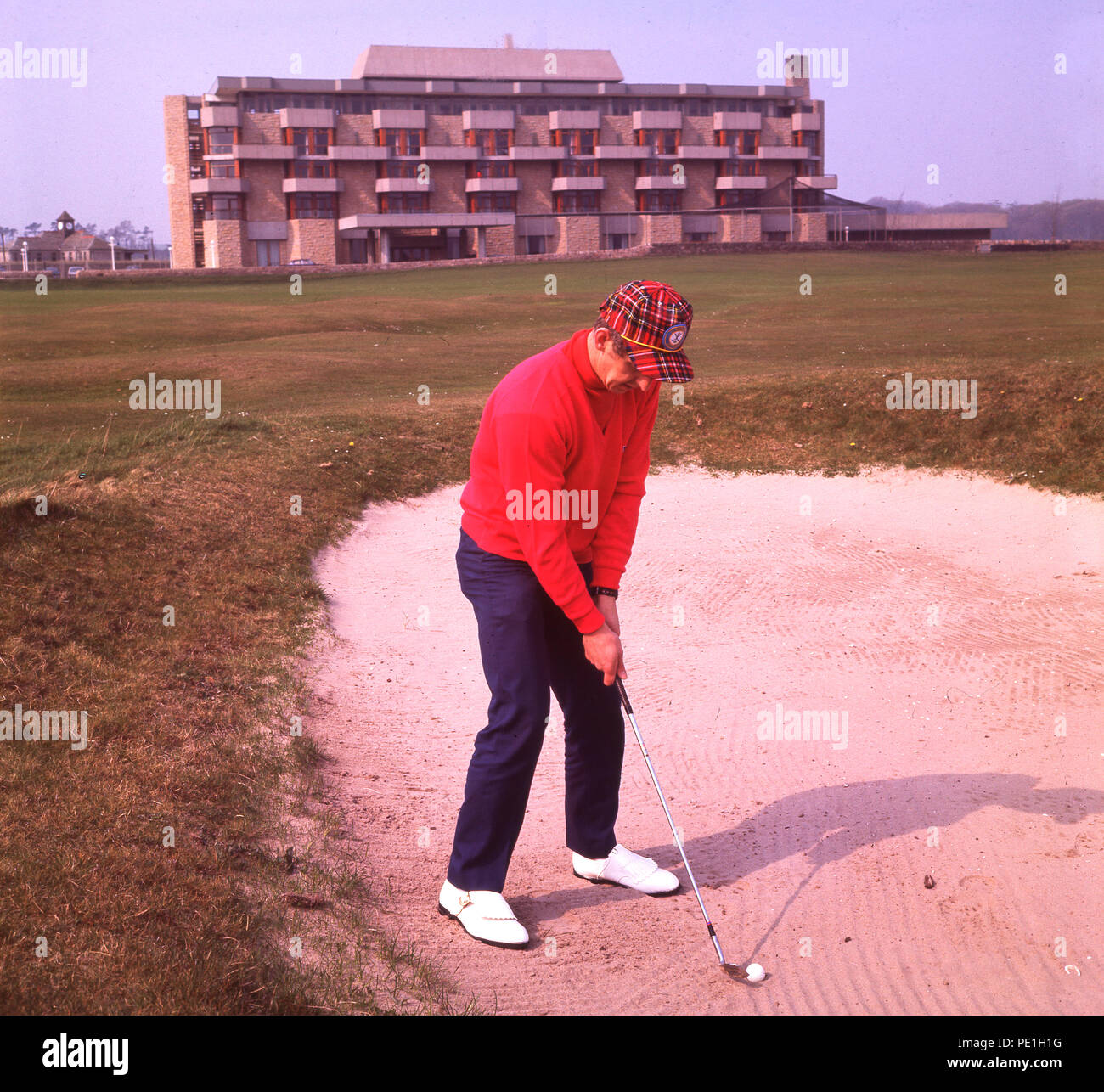 1968, historical, male golfer with tartan cap, playing out of a bunker on the old course at St Andrews golf links, with the newly built Old Course hotel in the background, St Andews, Fife, Scotland, UK. The unusual looking building was constructed by British Rail on the site of the old railway station and for such a prestigious piece of land, the hotel was a distinctly modest affair. Stock Photo