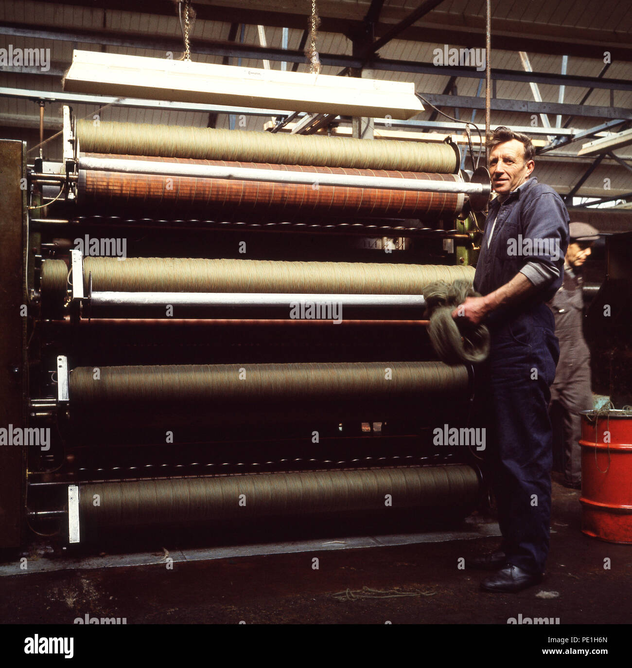 1960s, proud male worker standing by a carding machine at Harris Tweed, Stornoway, Lewis, Western Isles, Scottih Highlands, Scotland, UK. Carding is the process by which the individual wool fibres are straighened and sortted into separate fibres. Stock Photo
