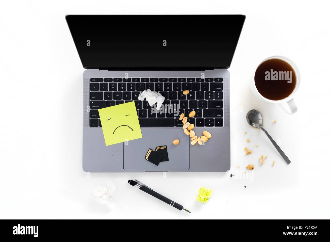 Top view of tabletop with messy dirty laptop, post it notes with sad face, nuts, memory cards, pen, spoon and coffee cup isolated on white background Stock Photo