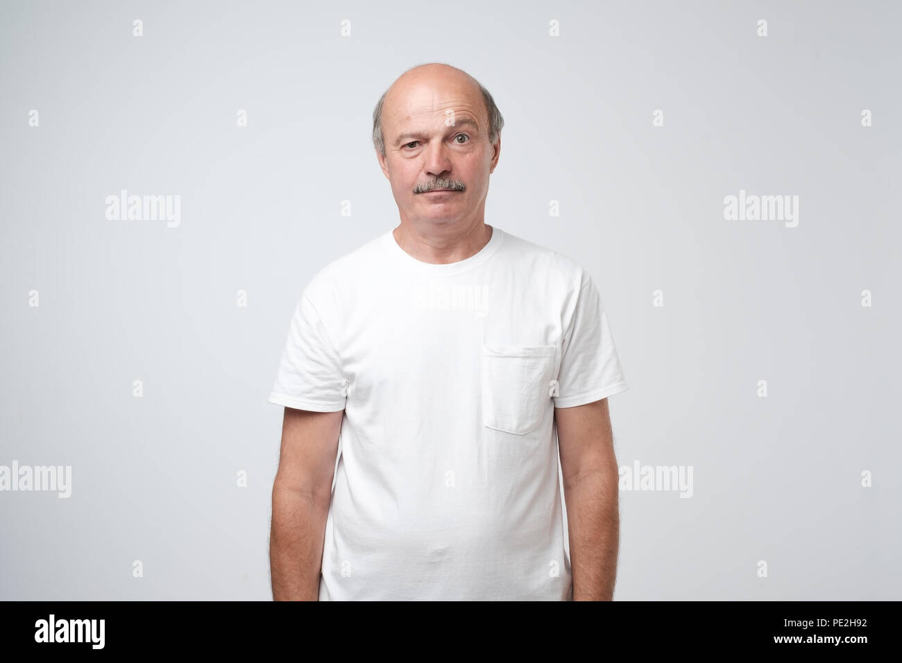 Elderly man emotions, portrait of serious senior caucasian man in white t-shirt looking at camera against gray wall Stock Photo