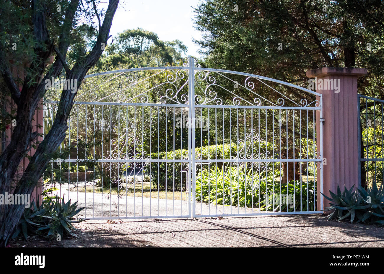 White wrought iron metal driveway entrance gates set in cement brick pillar with residential garden in background Stock Photo