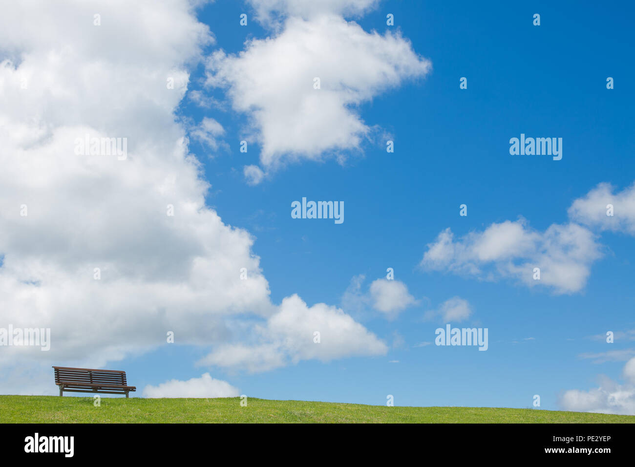 Very small bench in the distance on a grassy bank with a large blue sky and clouds in the background Stock Photo