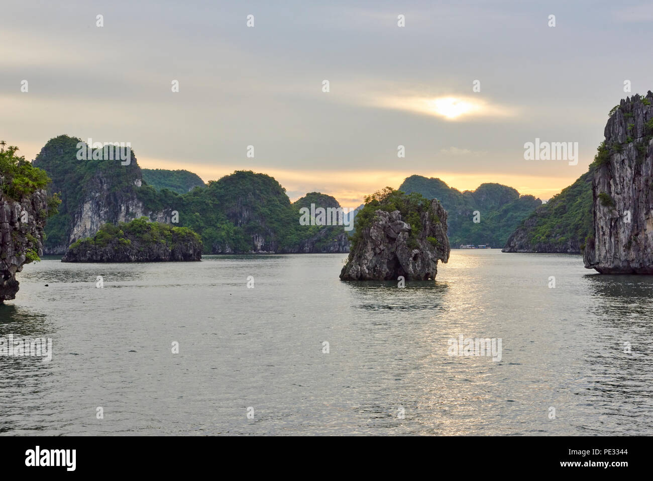 Sunset in Halong Bay, North Vietnam, with sun peaking through overcast sky and rocky islets in the mid ground. Stock Photo