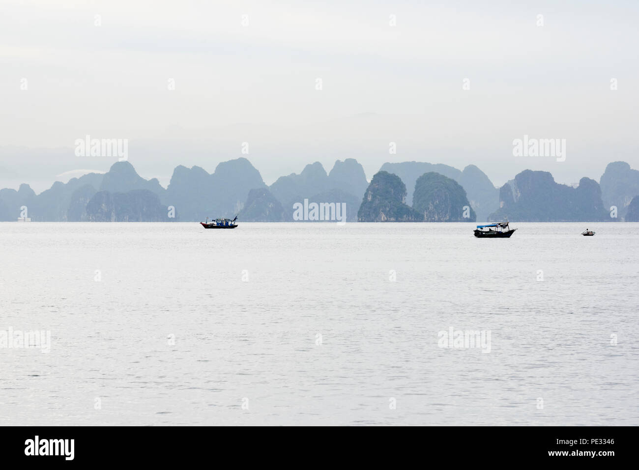 Quiet scenic seascape of Halong Bay, North Vietnam, with small fishing boats silhouetted against misty blue mountains in the background. Muted colours Stock Photo