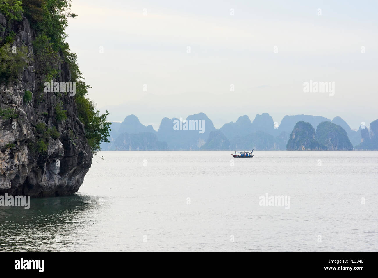 A small fishing boat in near silhouette at rest amongst range of islets in Halong Bay, North Vietnam, against misty blueish mountains in the backgroun Stock Photo