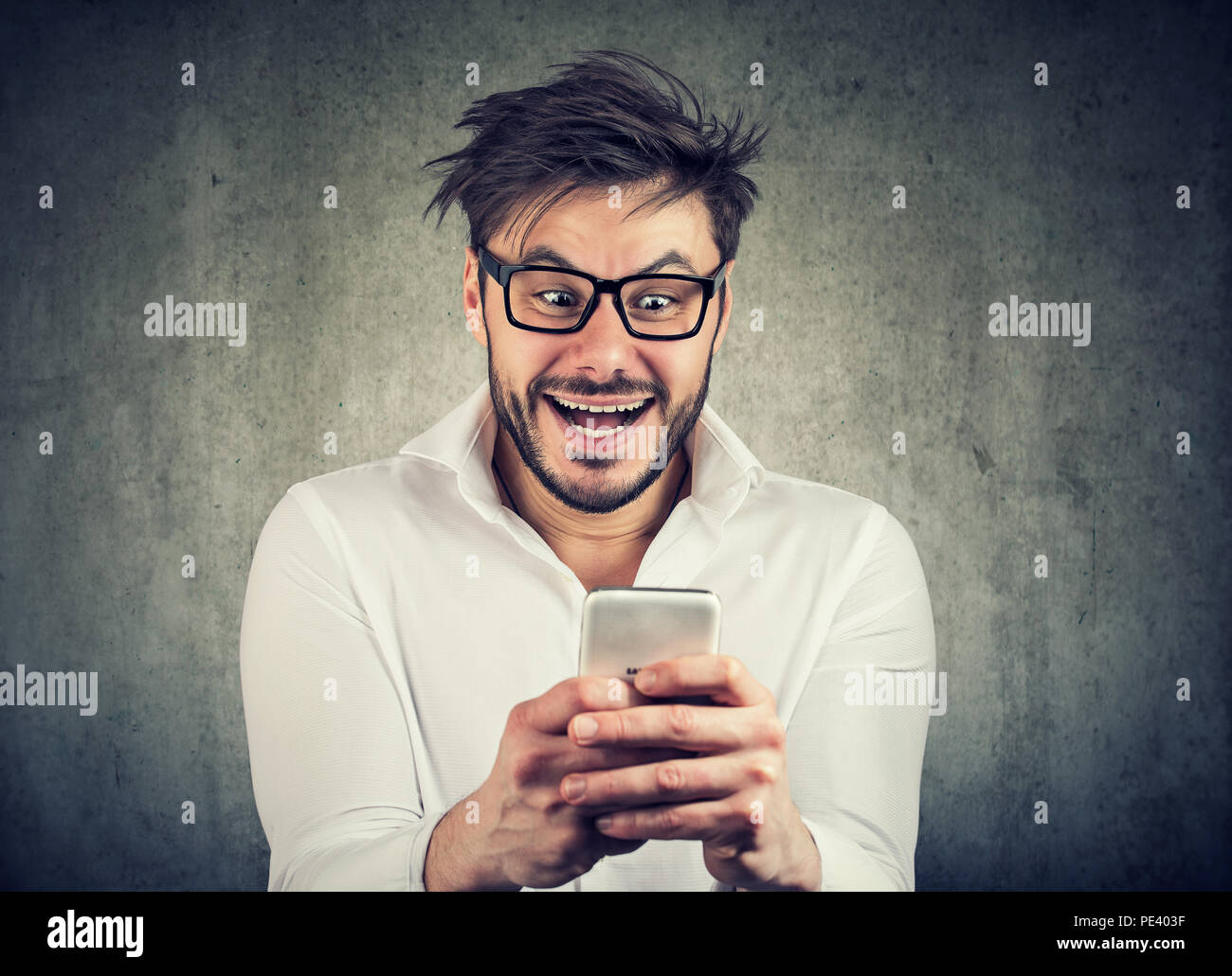 Thrilled and crazily looking young man in glasses and shirt watching news on smartphone standing against gray background Stock Photo