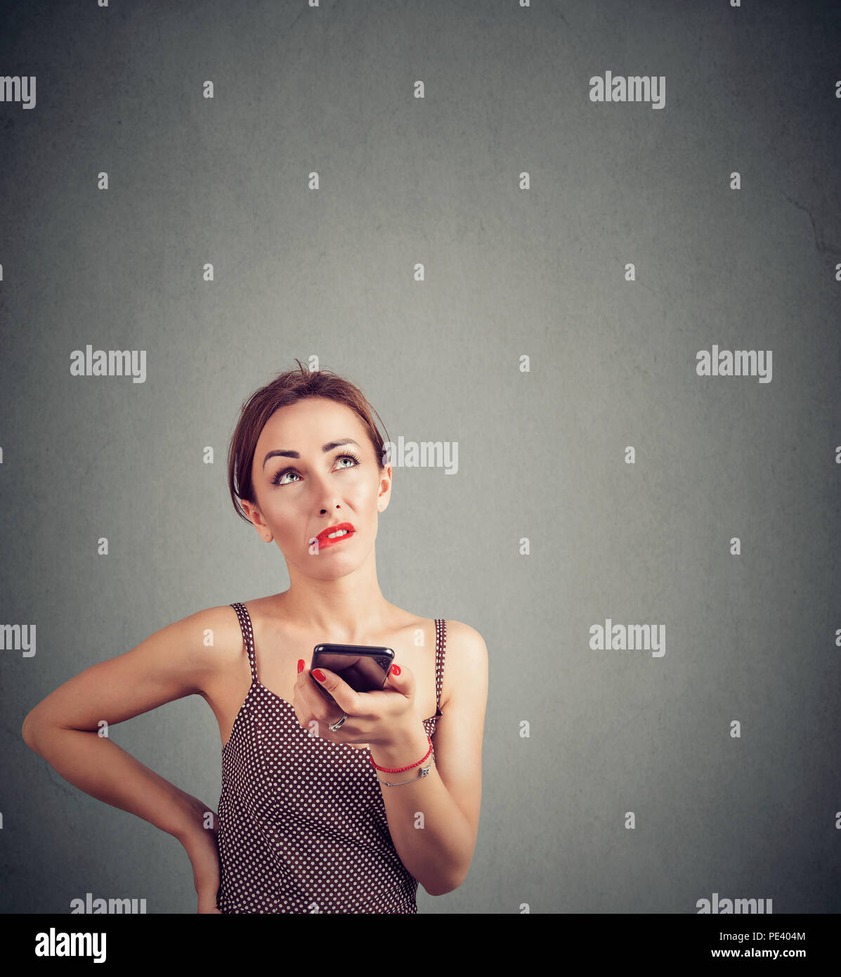 Young casual woman making grimace of irritation while speaking on smartphone and looking up on gray background Stock Photo