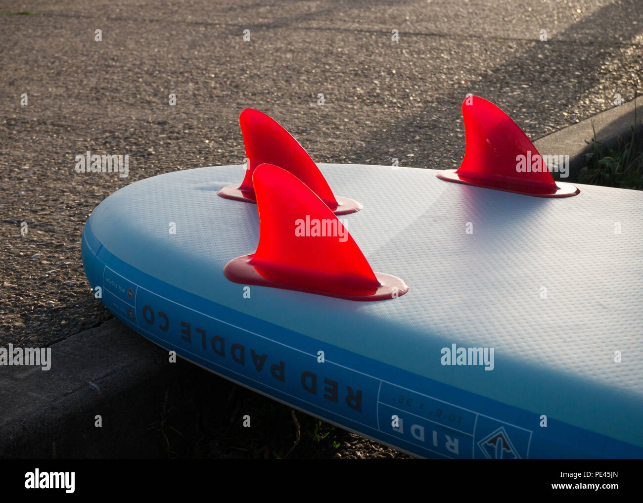 Colour close up of the red fins of a blue paddle board, lying upside down against a pavement