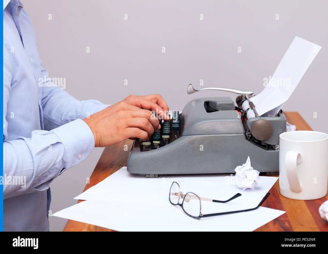 The man in the shirt typing on an old-fashioned typewriter. On the table are glasses and there is a mug. Neutral background Stock Photo