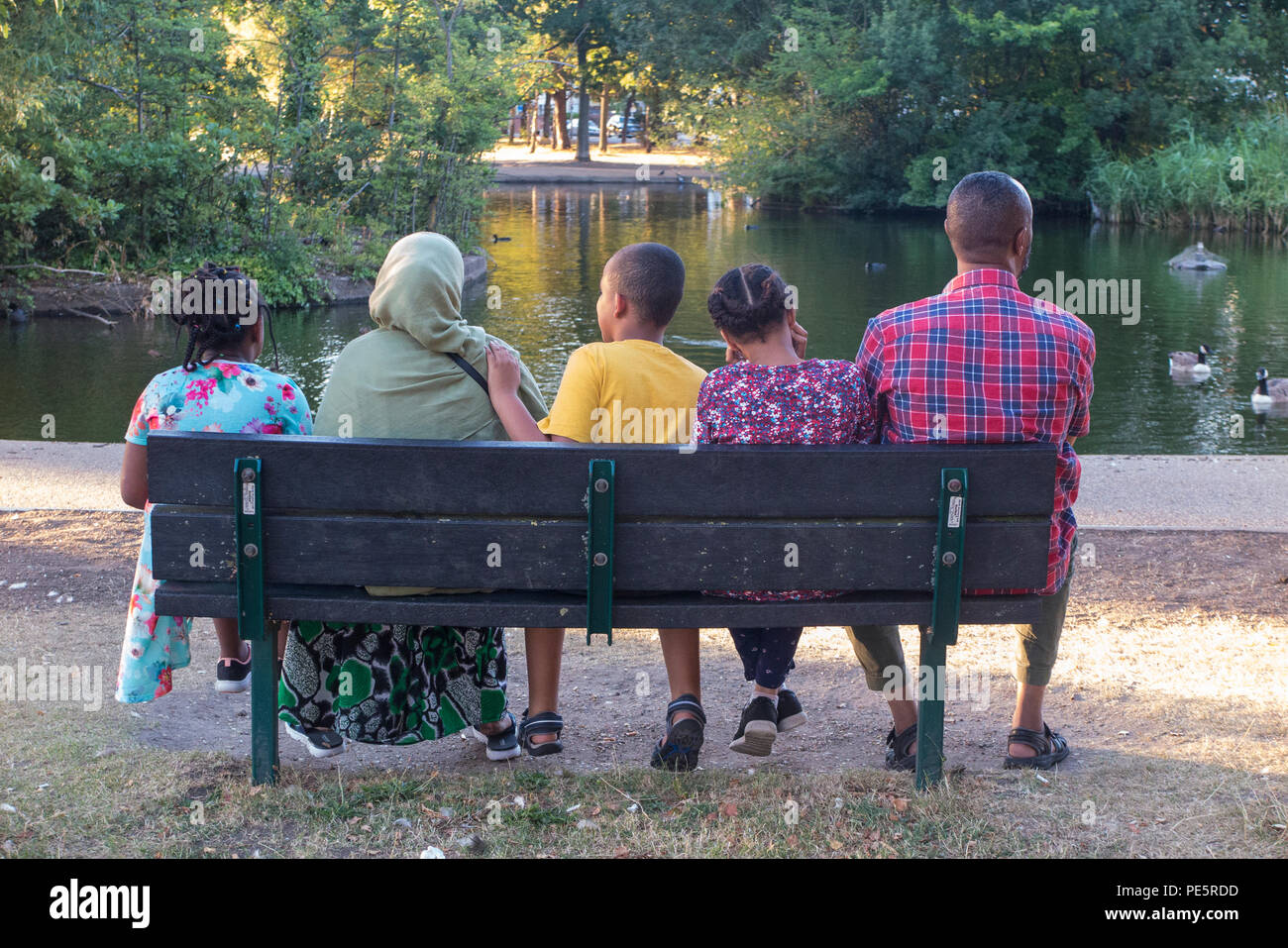 A muslim family enjoy the evening sunshine on a park bench in a London park Stock Photo
